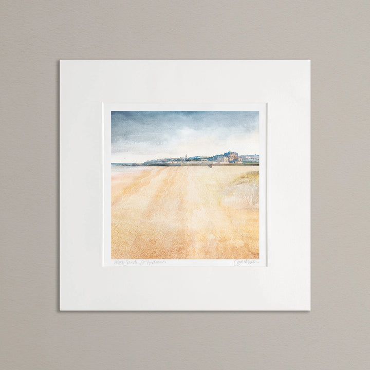 A mounted art print of West Sands, St Andrews, against a neutral grey background. The top 1/3 of the picture is the town shown as a view from the beach. The bottom part of the image is the beach. The sky is a muted blue grey. Overall the artwork is calming. There are 2 people walking in the distance on the beach