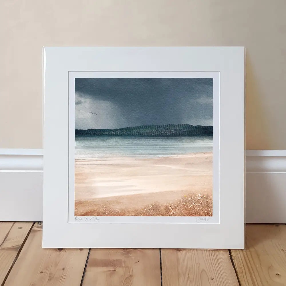 mounted print of beach scene with pinky peachy colours of sand and greeny blue sea with a dark rain cloud in the distance A wee bird is flying in the sky and migh be a seagull