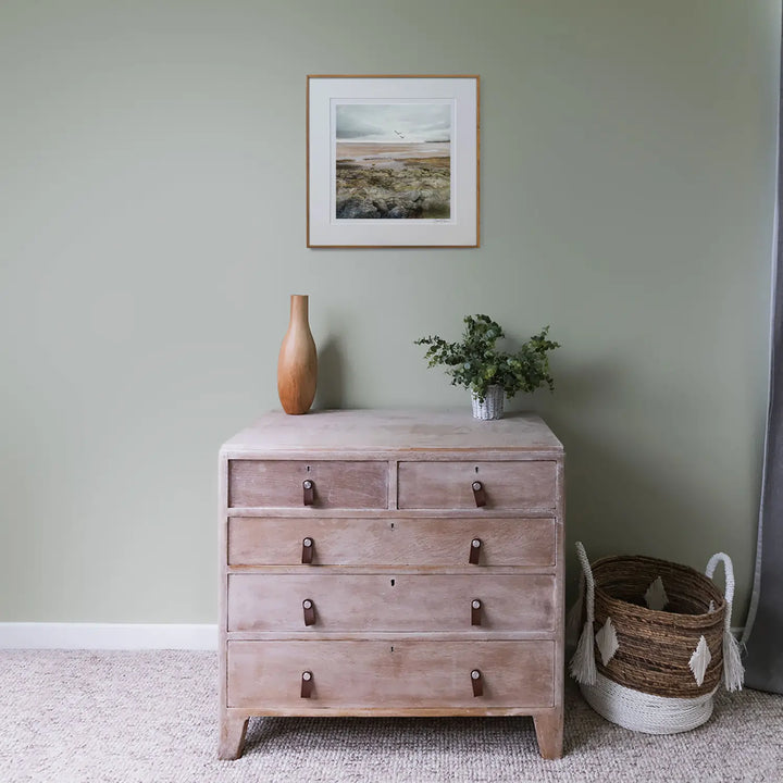 a green bedroom with a bleached oak chest of drawers with a framed print on the green wall behind. The decor is muted and relaxing.