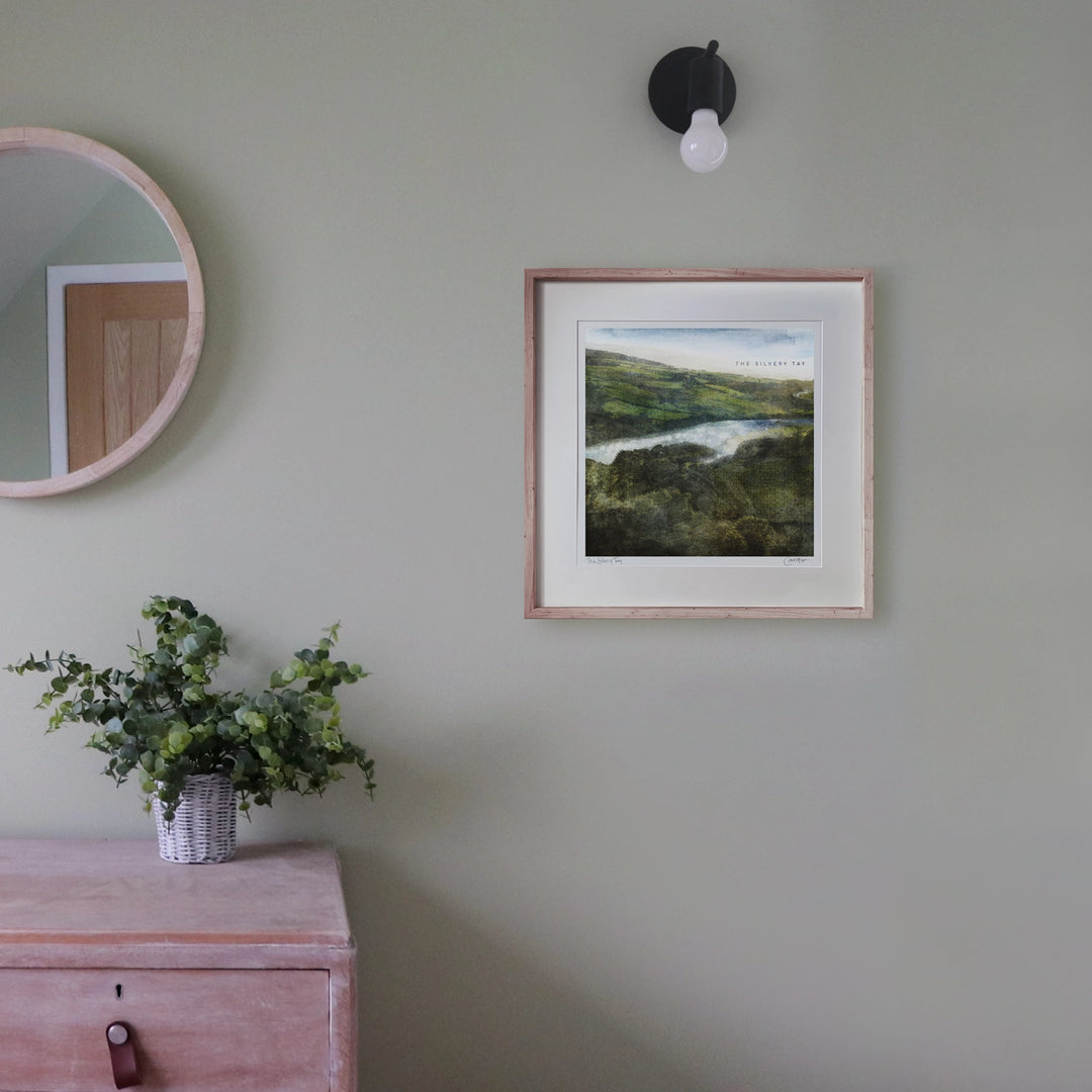 The Silvery tay is a mostly green coloured art print. the view is from near Kinoull Hill towards the Tay river. The fields have textures in them like knitwear. This shows the art print framed in an oak frame on a light gray green bedroom wall with a mirror and chest of drawers off to the side.