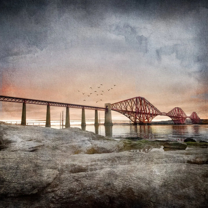 Queensferry Sunset closeup The picture is of the Forth Railway bridge with a sunset in the background and rocks in the foreground as it's from the South Queensferry side looking over to Fife. A flock of seagulls are flying over the bridge